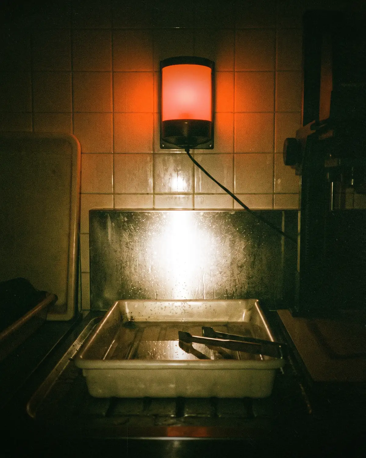 A darkroom under safelight. A developing tray on the bench, lit from behind.